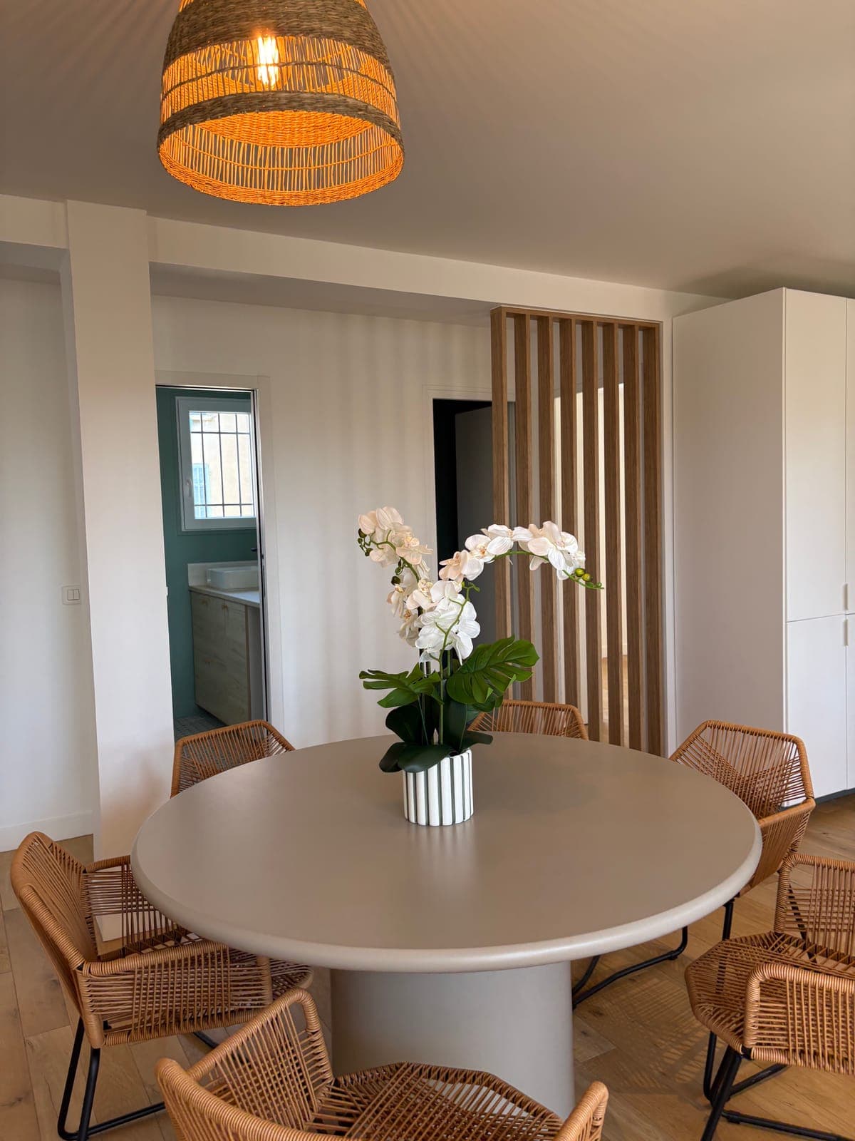 Round dining table and woven rattan chairs, in front of the wood-slat divider — Sanary-sur-Mer — two apartment furnishing project